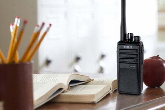 An RCA RDR2500 school two-way radio is set upright on a teacher's desk beside two textbooks, an apple, and a pencil holder.