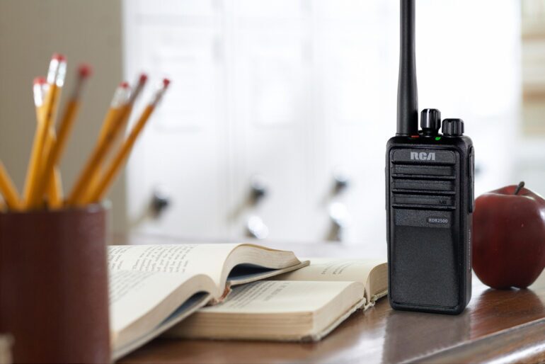An RCA RDR2500 school two-way radio is set upright on a teacher's desk beside two textbooks, an apple, and a pencil holder.